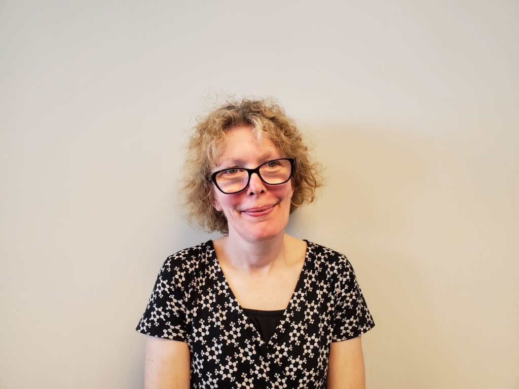 A photo of Becky Stewart. She is in her 50's with a slender frame and short curly grey/brown hair. She is wearing glasses, a black and white floral dress with short sleeves, and a black undershirt.