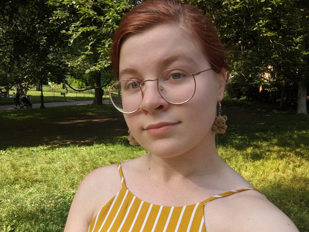 A photo of Hayley McGinniss. She is in her 20's with medium body type. She has straight hair up in a bun that is dyed dark red. She is wearing glasses, a white and yellow camisole, a septum nose ring, and teddy bear earrings.
