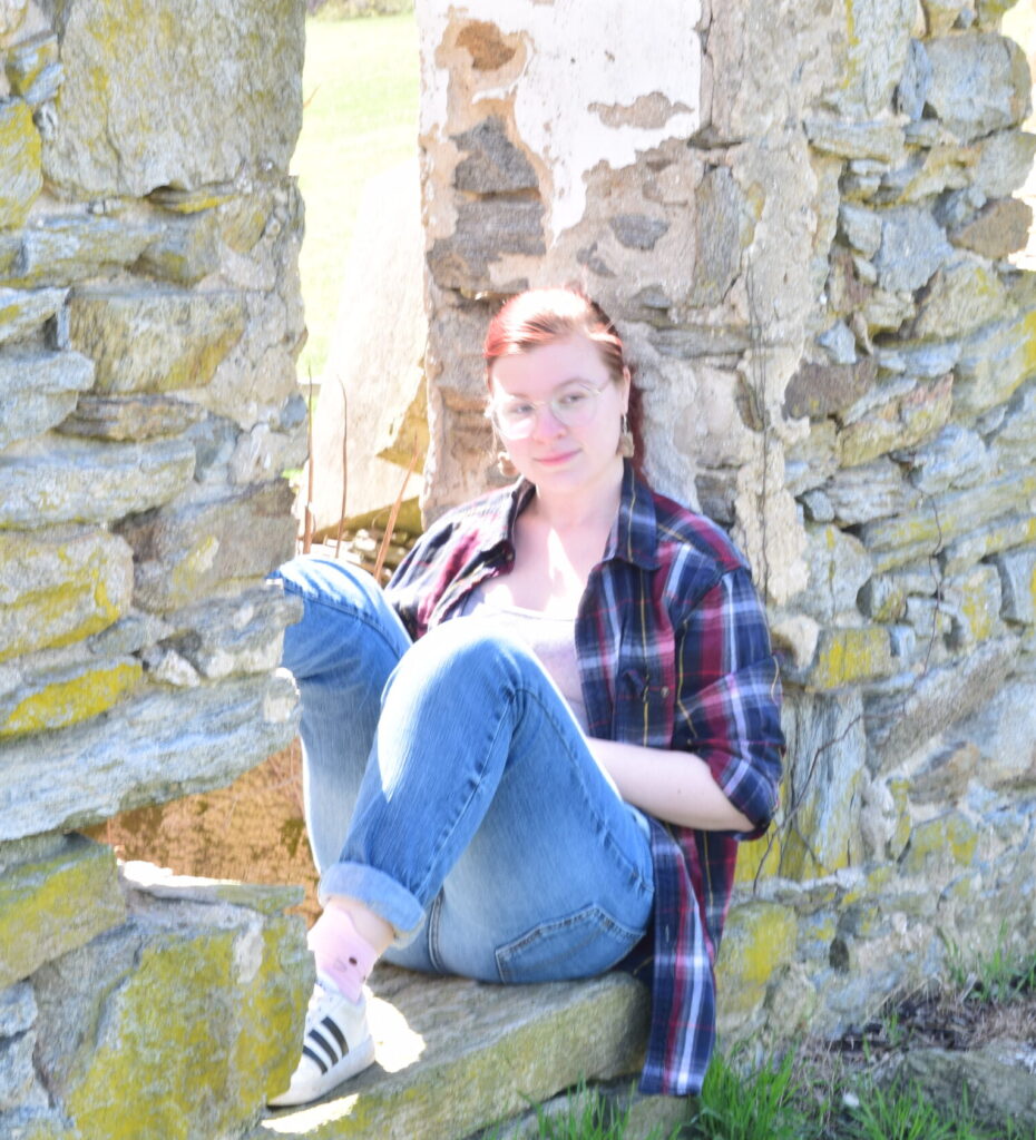 A casual photo of Hayley McGinniss, one of the editors. She is sitting in an old cobblestone windowsill. She is wearing glasses, a red and blue flannel, grey undershirt, blue jeans, white sneakers, and pink socks.