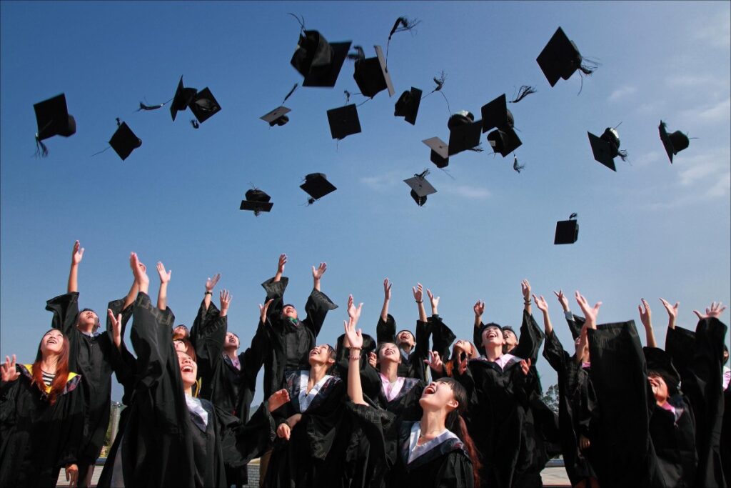 A photo of approximately 20 college students tossing their black graduation caps in the air. They are all dressed in black graduation gowns. 